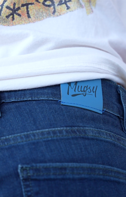Person wearing dark-wash Mugsy jeans with the signature blue Mugsy patch visible above the back pocket.